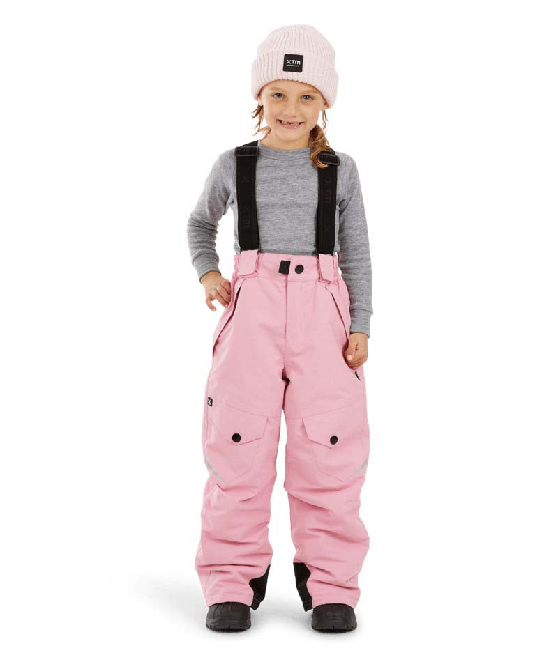 PlutoIIKidsPant-A young child wearing pink snow pants with black suspenders, a gray long-sleeve shirt, a light pink beanie, and black boots, standing against a plain white background.