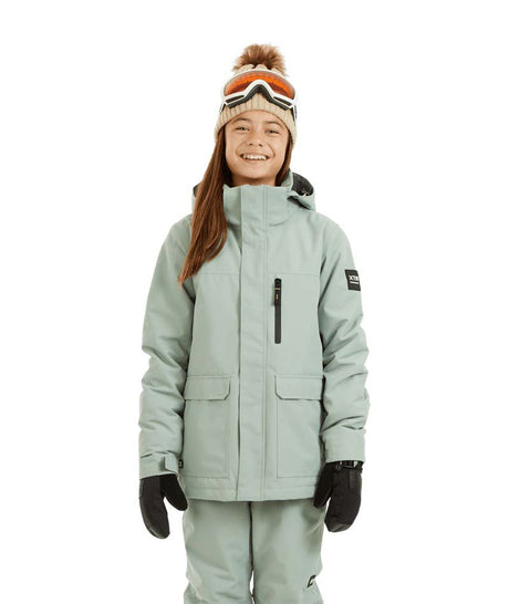 RumbleJacket- A smiling girl wearing a light sage green snowboard jacket, matching snow pants, black gloves, and a knit beanie with ski goggles, standing against a plain white background.