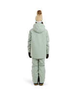RumbleJacket- A smiling girl wearing a light sage green snowboard jacket, matching snow pants, black gloves, and a knit beanie with ski goggles, standing against a plain white background.