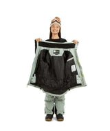 RumbleJacket- A smiling girl wearing a light sage green snowboard jacket, matching snow pants, black gloves, and a knit beanie with ski goggles, standing against a plain white background.