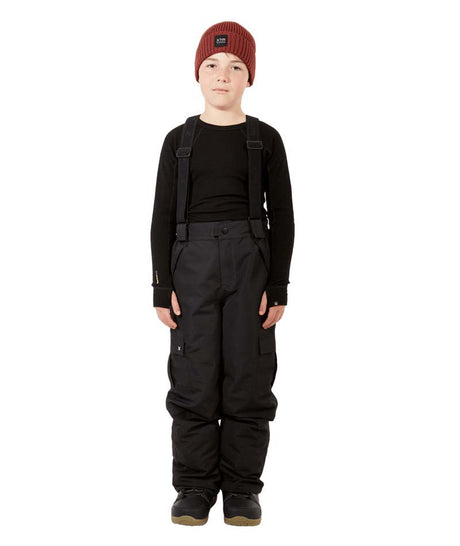 ScoobieIIPant-A child model wearing black snow bib overalls with suspenders, a black long-sleeve shirt, black snow boots, and a burgundy knit beanie, standing against a solid white background.
