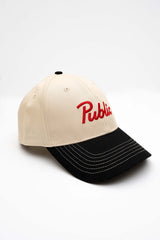 ScriptCap-A baseball cap with a cream-colored crown and a black curved brim, featuring the word 'Public' embroidered in red script on the front. The cap is photographed on a seamless white background.