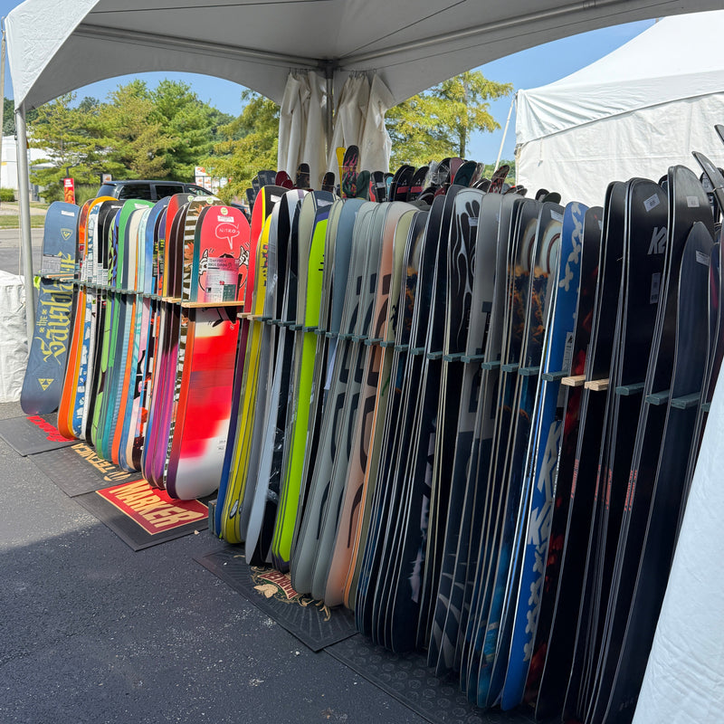 A row of colorful snowboards and skis leaning vertically under a white canopy tent at an outdoor market or event.