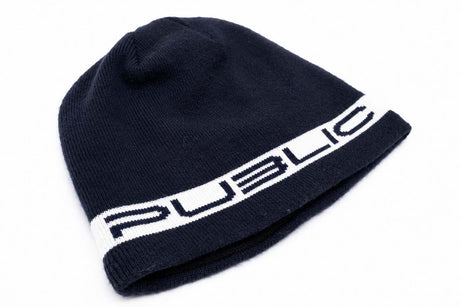 StretchLogoSkully- A dark navy blue knit beanie hat featuring a white horizontal band around the lower half with the word 'PUBLIC' in navy lettering, shown against a plain white 