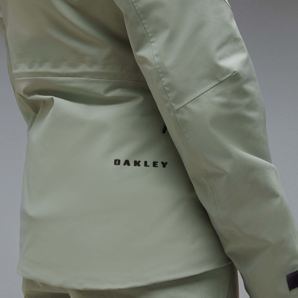 WCanopyInsulatedJacket-A light sage green hooded jacket with black zipper and drawstring details, displayed on a neutral gray background. The Oakley logo is visible on the left chest.