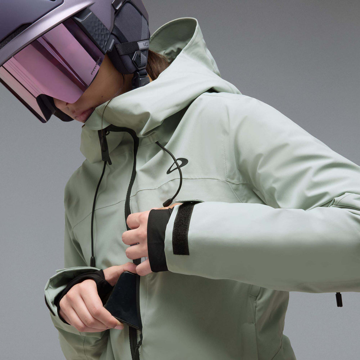 WCanopyInsulatedJacket-A light sage green hooded jacket with black zipper and drawstring details, displayed on a neutral gray background. The Oakley logo is visible on the left chest.