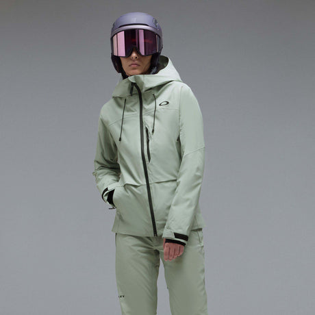 WCanopyInsulatedJacket-A light sage green hooded jacket with black zipper and drawstring details, displayed on a neutral gray background. The Oakley logo is visible on the left chest.