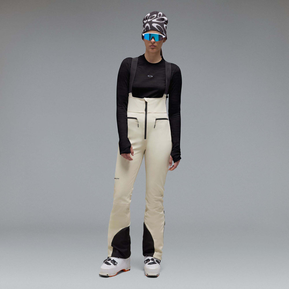 WIcicleSoftshellBib-A pair of ski bib pants in off-white with black suspenders, black zipper details, and black paneling at the lower legs, shown against a neutral gray gradient background.
