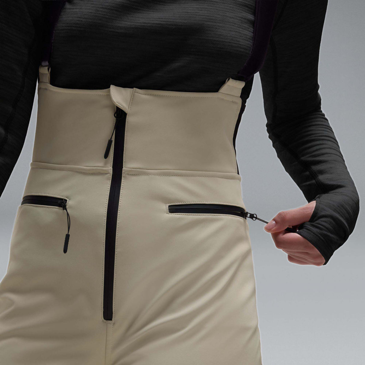 WIcicleSoftshellBib-A pair of ski bib pants in off-white with black suspenders, black zipper details, and black paneling at the lower legs, shown against a neutral gray gradient background.