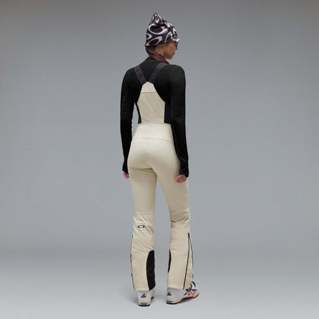 WIcicleSoftshellBib-A pair of ski bib pants in off-white with black suspenders, black zipper details, and black paneling at the lower legs, shown against a neutral gray gradient background.