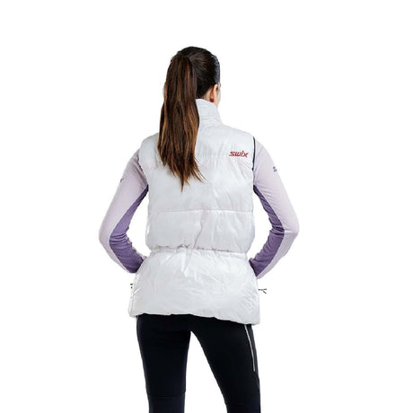 WMayenQuiltedPolyfillVest- A woman modeling a white sleeveless zip-front puffer vest with a colorful geometric pattern across the chest, black drawstring waist, and a red inner collar, worn over a light purple long-sleeve top and black pants against a white background.