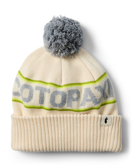 cumbre-2.0-beanie-A cream-colored knit beanie with a gray pom-pom, featuring a lime green stripe with 'COTOPAXI' knitted in gray across the middle and a ribbed cuff with a small black logo tag.