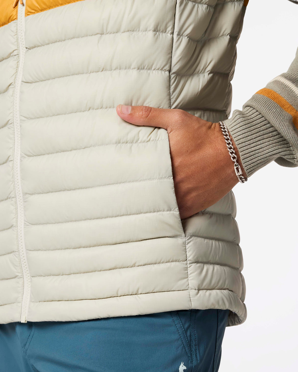 m-fuego-down-vest- A quilted puffer vest by Cotopaxi in a light beige color, featuring horizontal quilting, a full-zip front, and a mock neck collar. It has a distinctive color-blocked chest design with horizontal stripes of dark teal, rust orange, and golden yellow, with the Cotopaxi logo printed on the chest.