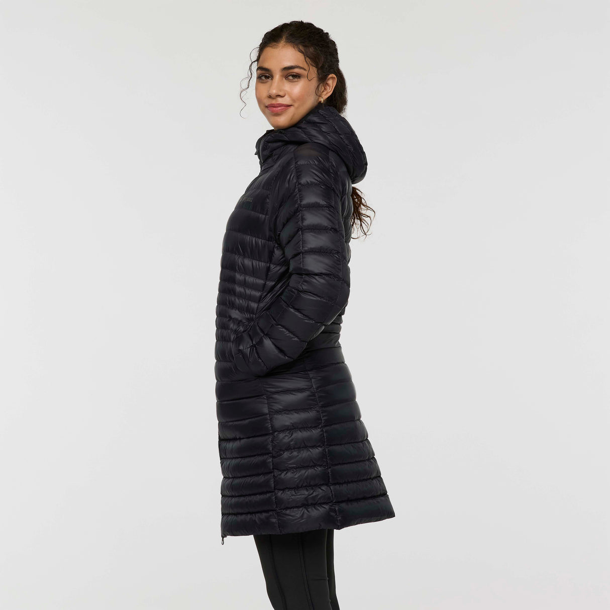 w-fuego-down-parka-A woman modeling a long, black quilted puffer jacket with a hood and a small embroidered 'Cotopaxi' logo on the left chest, set against a plain light gray studio background.